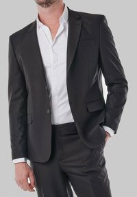 Veste de costume noire avec une texture lisse, design à une seule rangée de boutons, deux boutons et revers crantés, portée avec une chemise blanche à col en dessous.