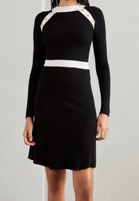Vestido negro de canalé con mangas largas, que presenta una cinturilla de canalé blanca y acentos en los hombros con botones dorados. Con cintura acampanada en la parte inferior.