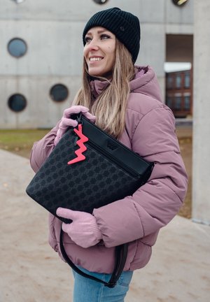 Black patterned handbag with a pink lightning bolt charm, held by a person in a pink puffer jacket and gloves, against a concrete backdrop.