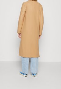 Beżowy, długi płaszcz o prostym kroju bez widocznych zapięć, w połączeniu z jasnoniebieskimi jeansami o szerokich nogawkach i niebieskimi tenisówkami.