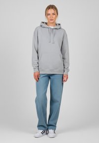 Felpa grigia chiara con cappuccio regolabile, tasca a marsupio e polsini a costine, abbinata a jeans a gamba larga blu e sneakers bianche.