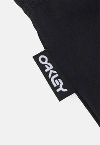 Svart stoff med teksturert overflate, som har en brodert hvit "Oakley"-logo på et rektangulært merke. Merket er sydd fast i kanten.