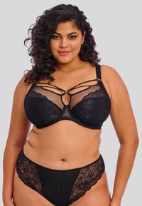 Reggiseno in pizzo nero con pannelli in rete, forma strutturata e cinturini decorativi incrociati. Accompagnato da un perizoma in pizzo coordinato.