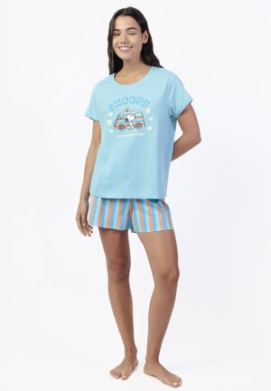 Joven sonriente, con camiseta azul claro con gráfico de Snoopy y pantalones cortos a rayas azul y naranja, de pie descalza sobre fondo blanco.