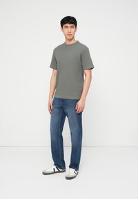 Jonge man voor een witte achtergrond, gekleed in een grijs gestructureerd T-shirt met korte mouwen, blauwe spijkerbroek en witte sneakers met zwarte strepen.