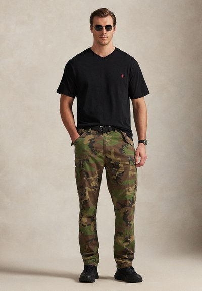 Άνδρας που φορά μαύρο T-shirt με V λαιμόκοψη, γυαλιά ηλίου, ρολόι, παντελόνι παραλλαγής τύπου cargo και μαύρα αθλητικά παπούτσια, στέκεται με τα χέρια στις τσέπες μπροστά σε μονόχρωμο φόντο.