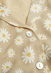 Beżowy kardigan o fakturowanym splocie, ozdobiony białymi stokrotkami z żółtymi środkami, wykończony okrągłym guzikiem z masy perłowej.