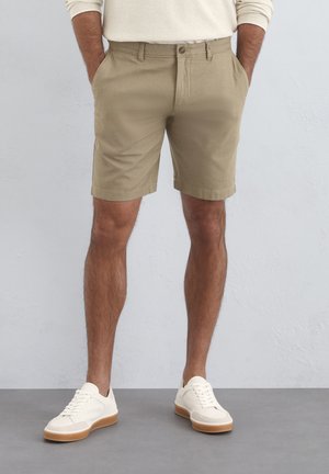 Tan cotton shorts with a straight fit, side pockets, belt loops, and a button closure, paired with white sneakers and minimal design details.