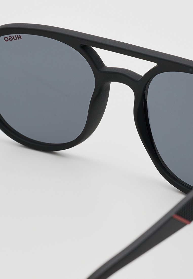 Schwarze Sonnenbrille mit matter Oberfläche, grauen Gläsern und einem eleganten, kantigen Design. Verfügt über einen strukturierten roten und schwarzen Akzent am Bügel.