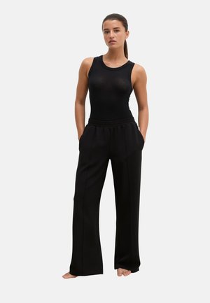 Femme aux cheveux foncés attachés en queue de cheval, porte un haut noir sans manches et un pantalon noir ample, debout pieds nus les mains dans les poches sur un fond uni.