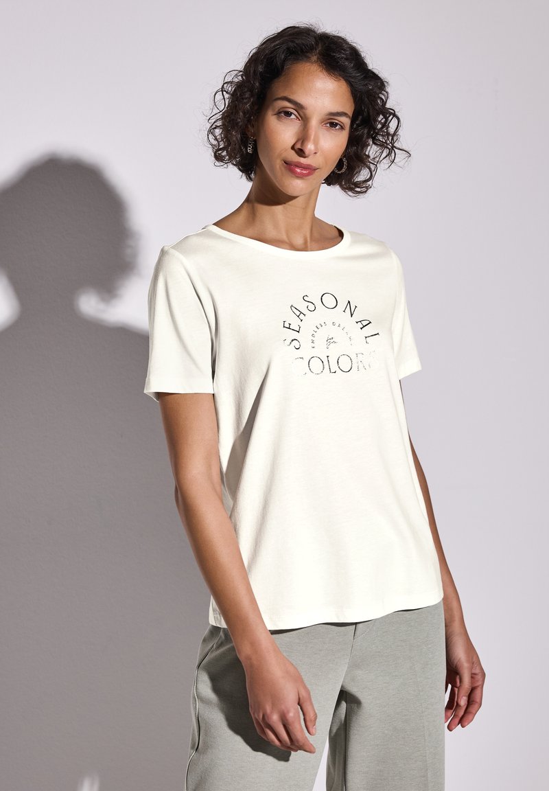Camiseta de color beige claro con mangas cortas, cuello redondeado y texto impreso que dice "COLORES DE TEMPORADA". Combinada con pantalones gris claro.