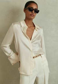 Camisa de satén crema con cuello clásico y mangas largas, combinada con pantalones beige ajustados y un cinturón delgado con un acento dorado.