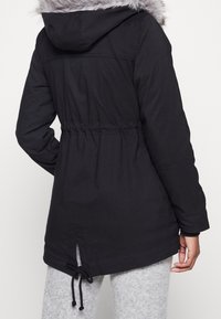 Svart parkas med en fuskpälsjacka, åtdragen midja, elastiska manschetter och dragsko i fållen. Har en mjuk och slät textur.