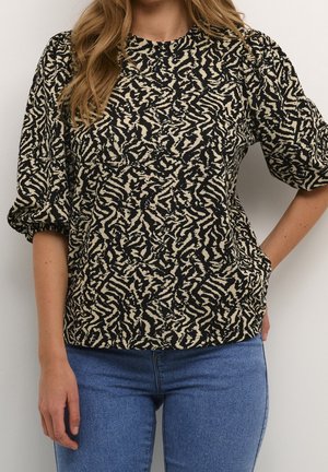 Donna che indossa una camicetta beige e nera con motivo astratto, maniche a sbuffo e jeans blu su uno sfondo semplice.