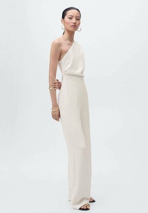 Mono de un solo hombro en color crema, con un corte relajado, piernas anchas y textura suave. La modelo lleva joyas doradas y sandalias negras.