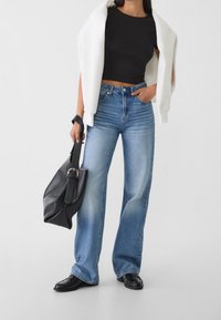 Mujer con pantalones vaqueros de pierna ancha azules, top corto negro, chaqueta blanca sobre los hombros, mocasines negros y sosteniendo un bolso negro.