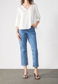 Vit blus med V-hals och trekvartsärmar, parat med ljusblå avkortade jeans. Modellen bär svarta sandaler med remmar.