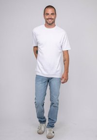 Homme souriant, portant un t-shirt blanc uni, un jean bleu clair, des baskets blanches et se tenant devant un fond gris neutre.