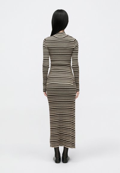 Vestido longo e ajustado com riscas verticais em bege e preto, com uma textura em canelado e gola alta. Usado com sapatos pretos.