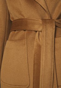 Detalle de un abrigo de lana marrón que muestra un bolsillo parche cosido y un cinturón de tela a juego atado a la cintura.