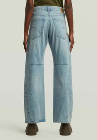 Persoon in lichtblauwe wijde jeans met achterzakken en bruine chunky laarzen, staand tegen een effen achtergrond.