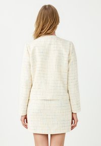 Vrouw in een crèmekleurige textuurjack en bijpassende rok met een subtiel blauw-geel ruitpatroon, staande met de rug naar ons gekeerd.