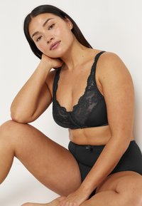 Musta pitsinen bralette, jossa on leveät olkaimet ja syvä V-pääntie, jossa on kukkakuviota, yhdistettynä korkeavyötäröisiin mustiin brief-pöksyihin, joissa on nauhaketju.