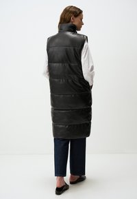Gilet sans manches noir matelassé avec col montant et finition brillante, porté sur une chemise blanche, associé à un jean foncé et des mocassins noirs.