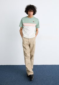 Lys grøn og hvid t-shirt med rosa vandrette striber, parret med beige bukser. Sorte sko fuldender outfittet. Ensfarvet baggrund.