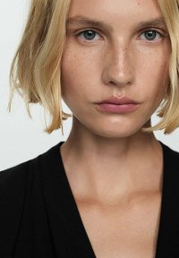 Närbild av en person med kort blont hår, ljus hy, blå ögon och naturlig sminkning. Bakgrunden är enkel vit; klädseln är svart.