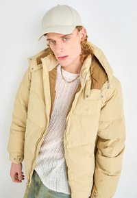 Beige puffer jacket with a hood, zip-front, and side pockets, layered over a white knit sweater. A light-colored cap completes the look.