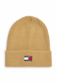 Gebreide beanie in lichtbruin met een omgeslagen ribgebreide rand. Bevat een merklabel met blauwe, witte en rode accenten. Zachte textuur.