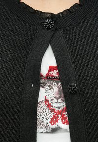 Mustast lõngast kootud kardigan, millel on ehtest nupud, mis on osaliselt lahti tehtud, paljastades valge särgi, millel on detailne leopard, kes kannab punast kübarat ja tikitud kaelakeed.