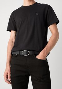 Czarny bawełniany t-shirt z krótkimi rękawami i okrągłym dekoltem, z dyskretnym logo na piersi. Połączony z czarnymi spodniami i srebrnym paskiem.