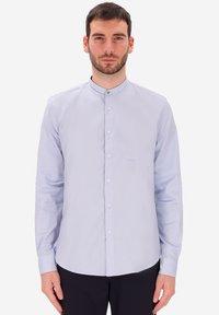 Uomo che indossa una camicia a maniche lunghe con colletto alla cinese di colore azzurro chiaro, con bottoni bianchi, in piedi contro uno sfondo bianco uniforme.