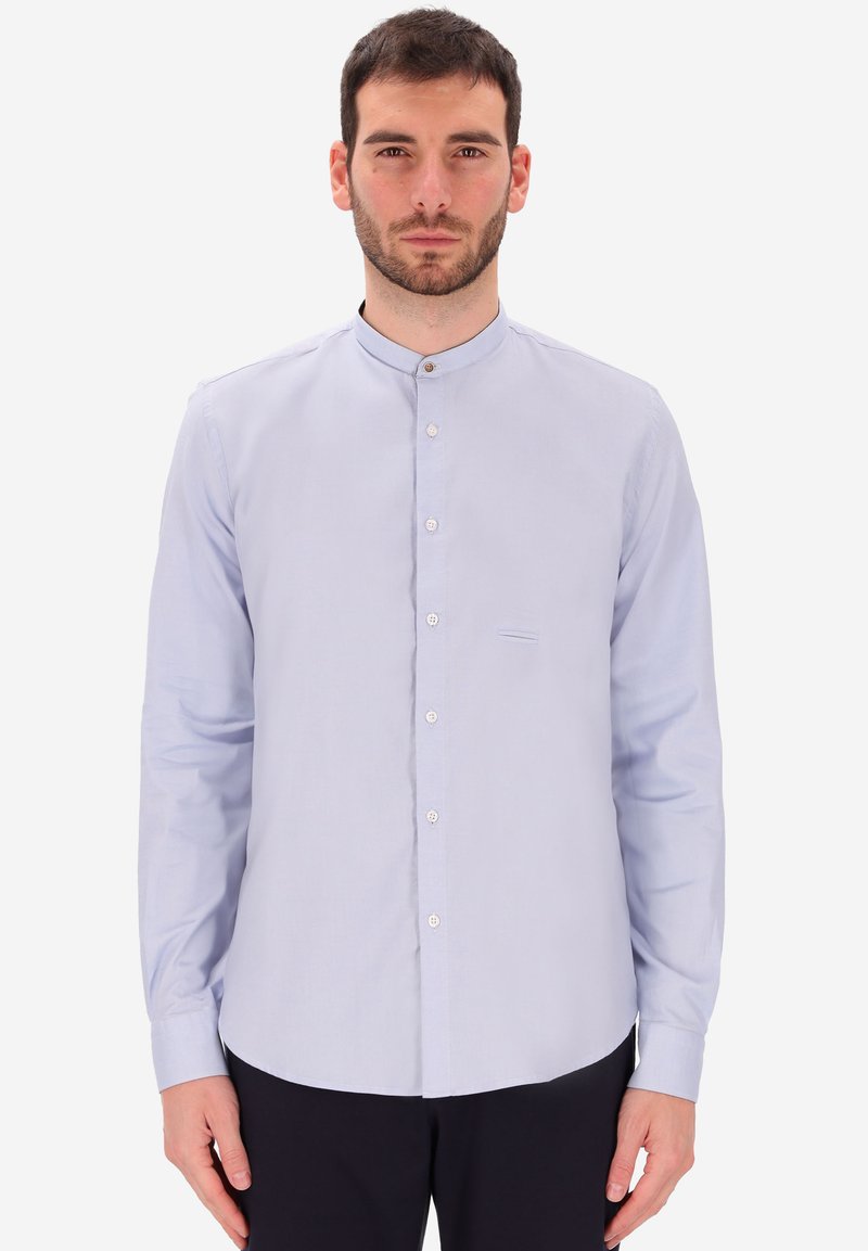 Uomo che indossa una camicia a maniche lunghe con colletto alla cinese di colore azzurro chiaro, con bottoni bianchi, in piedi contro uno sfondo bianco uniforme.