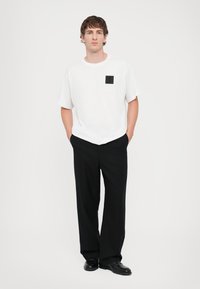 Hvit bomulls T-skjorte med en firkantet svart logo-lapp, kombinert med svarte vide bukser med vertikale pinstriper og svarte sko.