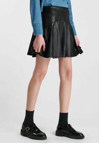 Zwarte faux leren uitlopende rok met een nauwsluitende tailleband en geplooid ontwerp. Draagt samen met zwarte schoenen en gestructureerde sokken, tegen een effen achtergrond.
