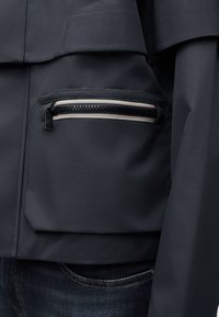 Σκούρο γκρι μπουφάν με μπροστινή τσέπη και λεπτομέρεια φερμουάρ με ρίγες. Το υλικό έχει λεία υφή και άνετη εφαρμογή.