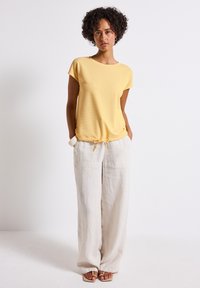 Žluté tričko s krátkým rukávem a strukturovaným designem, s nastavitelným stahovací páskem v pase, spárované s volnými světle béžovými lněnými kalhotami.