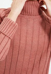 Rippschaliger Rollkragenpullover aus staubigem Rosé, mit hohem Kragen, langen Ärmeln und strukturiertem Stoff mit vertikalen Linien.