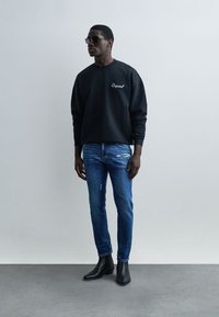 Man die een zwarte trui, blauwe versleten jeans, zwarte laarzen en een zonnebril draagt, leunend tegen een eenvoudige grijze muur en vloer.