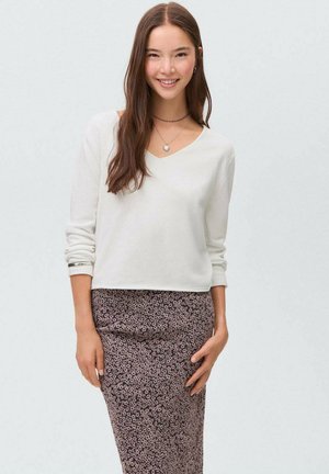 Weißer V-Ausschnitt-Pullover mit langen Ärmeln, kombiniert mit einem figurbetonten, floral gemusterten Rock in Brauntönen und Beige, getragen mit geschichteten Halsketten.