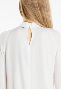 Weiße Bluse mit einem Schlüssellochverschluss auf der Rückseite und einem Knopf, aus glattem Stoff. Das Design umfasst subtile Schulternähte und eine lässige Passform.