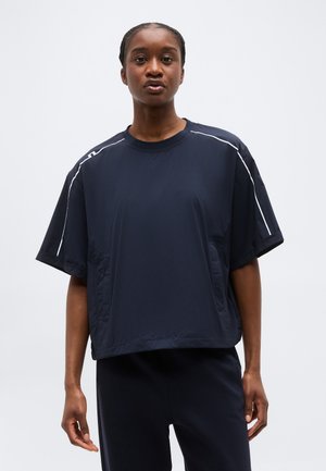 Γυναίκα με πλεκτά μαλλιά που φοράει άνετο σκούρο μπλε μπλουζάκι με κοντά μανίκια και λευκές ρίγες στους ώμους, συνοδευόμενο από παντελόνι σετ, μπροστά σε απλό φόντο.