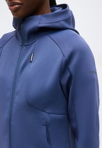 Κοντινό πλάνο ατόμου που φοράει μπλε φούτερ με φερμουάρ, υφασμένο με υφή και μικρή μαύρη ετικέτα λογότυπου στο στήθος και το μανίκι.