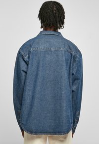 Denim krekls vidējā zilā krāsā, brīvs piegriezums, norādīta apkakle, garas piedurknes ar pogām, un taisna apakšmalu. Aizmugurējā skata attēlo šuvju detaļas.