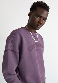 Lilla sweatshirt med rund hals, detaljer med frynseterm på ermene og brodert tekst. Bært med et sølvkjede.