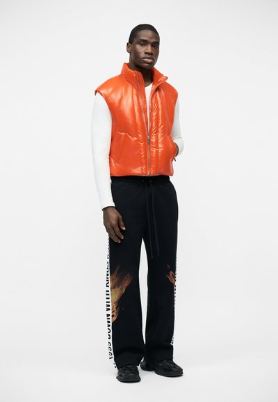 Άνδρας που φοράει λαμπερό πορτοκαλί αμάνικο μπουφάν τύπου puffer, λευκό πουλόβερ με μακριά μανίκια, μαύρη φόρμα με σχέδια φλόγας και μαύρα αθλητικά παπούτσια, όρθιος.