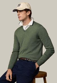 Uomo con cappellino beige, camicia bianca con colletto sotto maglione verde a scollo a V, pantaloni blu navy, cintura marrone, seduto su uno sgabello di legno, che guarda a sinistra.